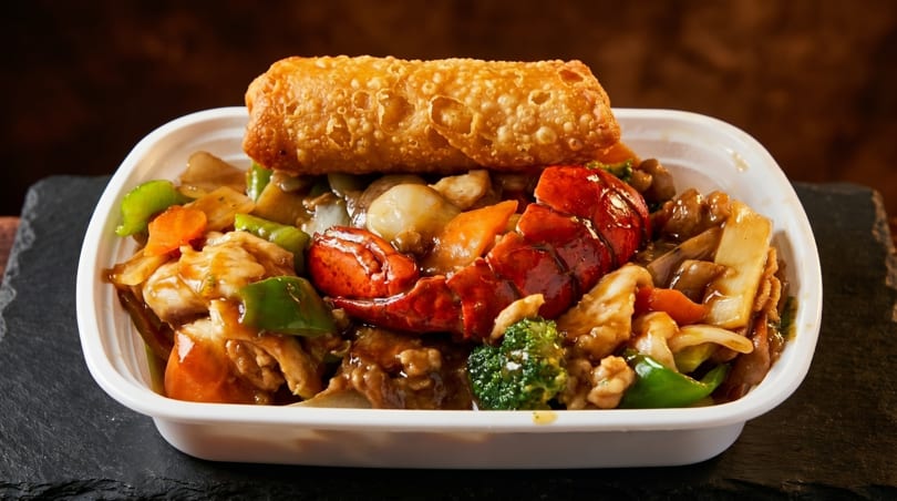 Lobster and chicken stir-fry with vegetables and an egg roll in a white takeout container.